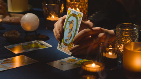 Close-Up-Of-Woman-Giving-Tarot-Card-Reading-On-Candlelit-Table-Holding-The-World-Card